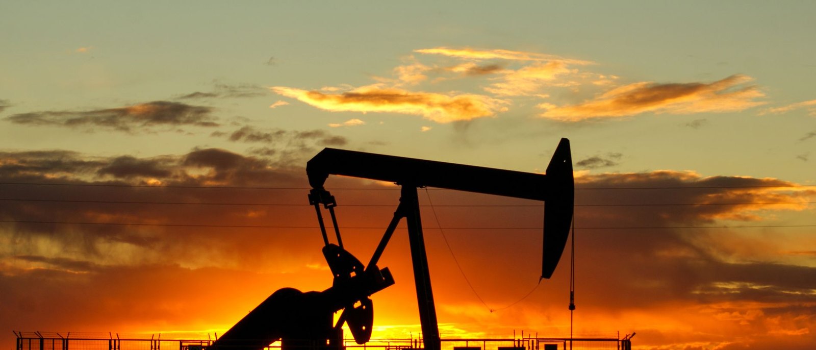 Pump jack silhouetted against a deep orange and gold sunset sky over a fenced oilfield production site
